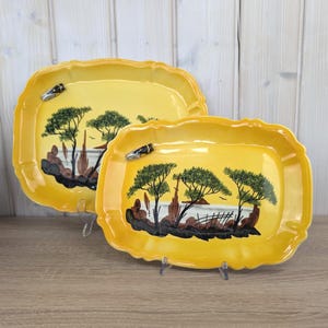 Puede incluir: Dos fuentes de cerámica amarillas con bordes festoneados. Cada fuente presenta un paisaje con árboles, rocas y una masa de agua. Las fuentes tienen un acento decorativo de insecto en el borde.