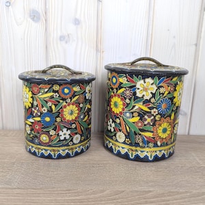 May include: Two vintage metal canisters with lids. The canisters are cylindrical and decorated with a floral pattern in yellow, red, blue, and white on a black background. Each canister has a handle on the lid.