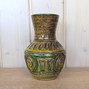 Könnte beinhalten: Eine Keramikvase mit bauchigem Körper und ausgestelltem Hals, verziert mit einem strukturierten Muster aus erhabenen vertikalen Rippen und kreisförmigen Motiven in Olivgrün, Senfgelb und Dunkelbraun mit glänzender Glasuroberfläche.