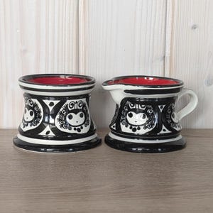 May include: Two ceramic pieces, a mug and a small pitcher, feature a black and white owl design. The interior of each piece is red. The owl design is repeated around the body of each piece. The mug has a cylindrical shape, while the pitcher has a handle and spout.
