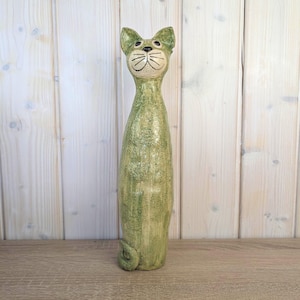 Puede incluir: Una figura de gato de cerámica verde alta con un diseño caprichoso. El gato tiene un cuerpo largo y delgado, una cara sonriente y una cola enroscada. La figura está pintada en tonos verdes y tiene una superficie texturizada.
