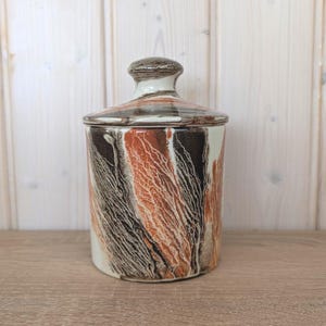 Op de afbeelding: Een keramische pot met deksel, met een ontwerp van verticale, getextureerde lijnen in bruin, zwart en wit. De pot heeft een cilindrische vorm en een afgeronde deksel met een kleine knop. De algehele esthetiek is rustiek en aards.