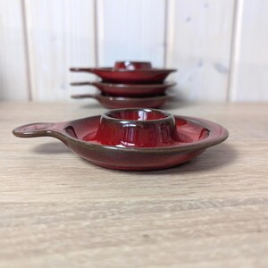 May include: A collection of six red ceramic egg cups with a dark brown rim and handle. The egg cups have a glossy finish and are arranged on a light wooden surface. Each cup has a circular base with a central egg holder.