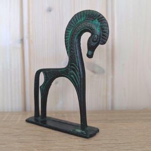 May include: A dark green and black metal horse sculpture with an arched neck and stylized details. The sculpture has a rectangular base and stands upright. The horse's mane and body have green accents, adding contrast to the dark metal.