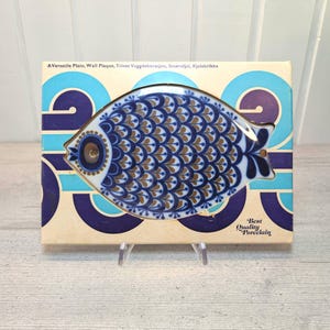 Könnte beinhalten: Ein fischförmiger Porzellanteller mit blau-braunem Schuppenmuster, präsentiert in einer cremefarbenen Schachtel mit blauen und violetten geometrischen Wellenmustern. Die Verpackung enthält den Text A Versatile Plate, Wall Plaque, Trivet und Best Quality Porcelain.