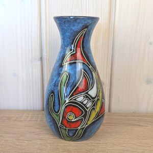 Può includere: Un vaso di ceramica blu con un disegno di pesce. Il vaso presenta una vivace illustrazione di pesce in rosso, giallo e bianco, delineata in nero, su uno sfondo blu. Sono presenti anche forme verdi simili ad alghe. Il vaso ha una parte superiore svasata.