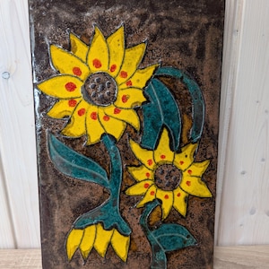 Puede incluir: Azulejo cerámico rectangular con diseño de girasoles. Los girasoles tienen pétalos amarillos con puntos rojos y centros marrones. Las hojas y los tallos son turquesa sobre un fondo marrón. Una pieza decorativa.