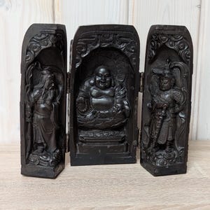 Pode incluir: Um biombo de madeira escura com figuras esculpidas. O painel central retrata um Buda sentado, ladeado por painéis com figuras de guerreiros. O biombo é intrincadamente esculpido com desenhos detalhados e tem uma construção articulada.