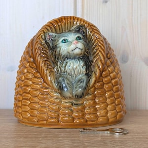 May include: A ceramic cat figurine nestled in a woven basket. The cat has green eyes and a pink nose. The basket is a warm brown color with a textured surface. A silver key rests on the wooden surface.