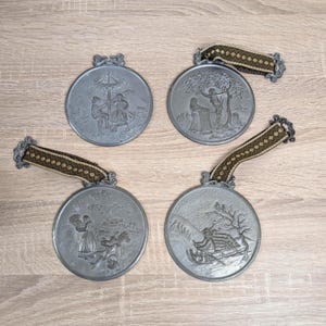 May include: Four antique pewter-coloured medallions with embossed scenes and decorative ribbon hangers. Each circular medallion depicts a different pastoral scene, including figures and landscapes. The ribbons are brown and green.