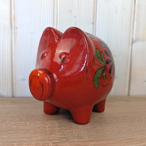 Könnte beinhalten: Ein glänzendes rotes Keramik-Sparschwein mit detaillierten grünen und schwarzen Blumenmustern. Das Sparschwein hat eine runde Schnauze, kleine Ohren und vier kurze Beine. Der Hintergrund ist helles Holz.