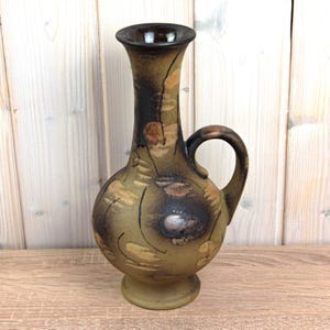 May include: A decorative ceramic vase with a handle. The vase has a bulbous body and a slender neck, with a dark brown rim. The exterior features a blend of brown, tan, and black hues, with a textured, painted design. The vase stands on a small, round base.
