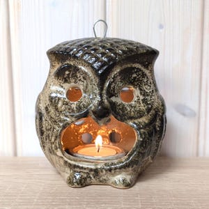 May include: A ceramic owl-shaped candle holder with a lit tea light inside. The owl is dark brown and grey with textured details. It has large eye openings and a small loop for hanging.