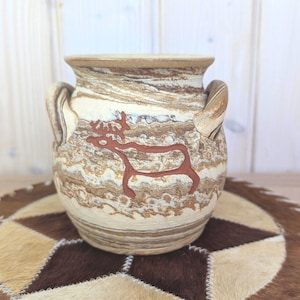 May include: A ceramic vase with a beige and brown marbled design and two handles. A red deer figure is painted on the front. The vase sits on a brown and beige patterned surface.