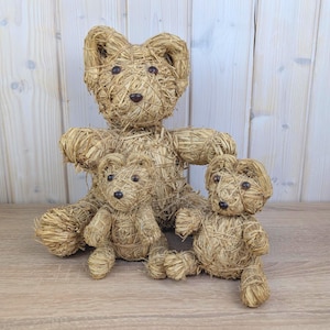 Pode incluir: Três ursos de pelúcia de palha de tamanhos variados. O urso maior está no fundo, com dois ursos menores na frente. Cada urso tem olhos de botão marrom escuro e um nariz marrom escuro. Os ursos são feitos de palha tecida.
