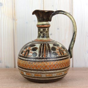 Może przedstawiać: Ceramiczny dzban z zaokrąglonym korpusem i uchwytem. Dzbanek ma ciemnobrązowy dzióbek i uchwyt, z dekoracyjnym wzorem pomarańczowych, czarnych i kremowych kształtów geometrycznych i motywów kwiatowych. Podstawa ma teksturowany, diamentowy wzór.
