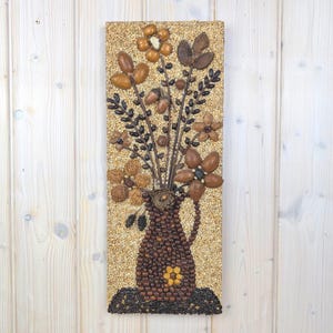 Puede incluir: Una obra de arte rectangular que presenta un jarrón lleno de un arreglo floral, todo hecho de varias semillas y nueces. El jarrón es marrón y el fondo es de color beige claro. Las flores y las hojas son en tonos marrones y beige.