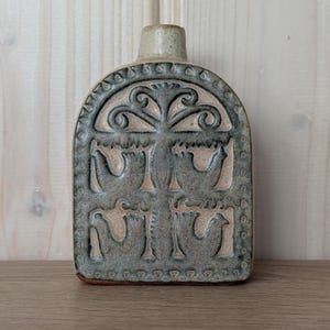 Pode incluir: Garrafa retangular de cerâmica cinza com gargalo e base bege. A garrafa apresenta um design em relevo de pássaros e padrões florais. O design é emoldurado por uma borda arqueada. A garrafa é exibida em uma superfície de madeira.