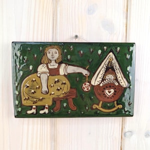 May include: A rectangular green ceramic wall plaque featuring a hand-painted illustration of a person in a yellow skirt sitting on a stool, holding a small round ornament near a brown rocking cradle with a heart design and a white canopy.