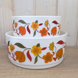 May include: Two white ceramic bowls stacked, featuring a floral pattern. The design includes orange, yellow, and red flowers with green leaves. The bowls have a rounded shape and are likely for serving or display.