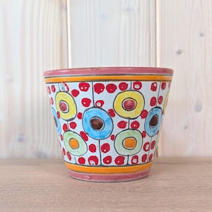 Może przedstawiać: Ceramiczna doniczka z białym tłem, ozdobiona powtarzającym się wzorem czerwonych kropek oraz kolorowych kół w odcieniach żółci, błękitu i zieleni. Krawędź i podstawa są podkreślone poziomymi pomarańczowymi i różowymi pasami, tworząc żywy wzór.