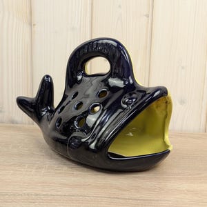 Könnte beinhalten: Ein dekoratives Objekt in Form eines Keramikwals. Der Wal ist hauptsächlich marineblau mit gelbem Innenraum und Maul. Er hat einen Griff oben und mehrere kreisförmige Löcher. Das Objekt steht auf einer Holzoberfläche.