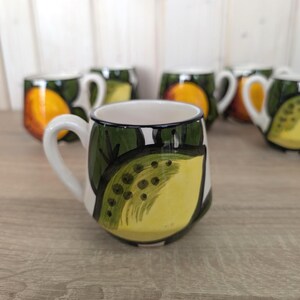 Könnte beinhalten: Weiße Keramiktasse mit einem bemalten Design einer gelben Zitronenscheibe und grünen Blättern. Die Tasse hat einen weißen Henkel und einen dunkelgrünen Rand. Im Hintergrund sind weitere Tassen mit Orangenfruchtmustern zu sehen.