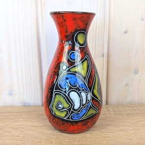 May include: A tall, ceramic vase with a vibrant red background and a fish design. The fish is outlined in black with yellow, blue, and white accents. The vase has a glossy finish and a narrow neck.