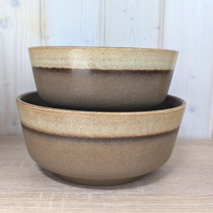May include: Two stacked ceramic bowls. The bowls have a beige rim, a dark brown band, and a medium brown base. The bowls are sitting on a light wood surface, with a light wood background.