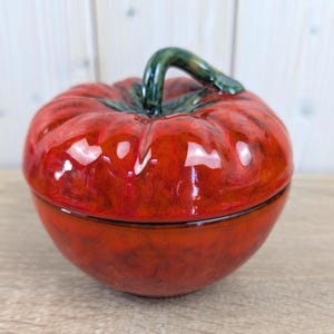 May include: A ceramic tomato-shaped container with a glossy red finish. The lid features a green stem, and the container has a textured surface. This decorative piece is likely used for storage or display.