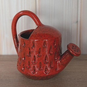 Può includere: Un annaffiatoio in ceramica rossa con un manico grande e un beccuccio con fori multipli. L'annaffiatoio ha una superficie strutturata con un motivo a goccia. Il manico è arcuato e il beccuccio è angolato.