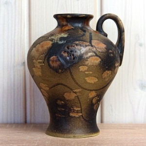 Könnte beinhalten: Eine Keramikvase mit dunkelbrauner und hellbrauner, gesprenkelter Glasur. Die Vase hat einen abgerundeten Körper, einen schmalen Hals und einen gebogenen Henkel. Die Oberfläche weist abstrakte Muster und Texturen auf.