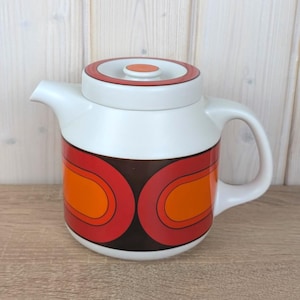 Peut inclure: Théière en céramique blanche avec couvercle et anse. La théière présente un design rétro avec une base marron et des formes incurvées rouges et oranges. Le couvercle a un anneau rouge et orange avec un petit cercle orange au centre.