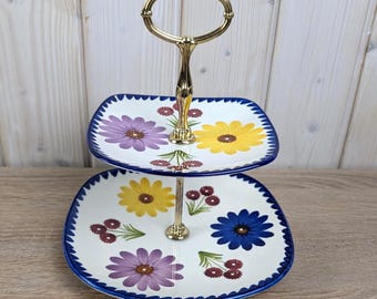 May include: Two-tiered serving stand with a gold-tone handle. The white plates have a blue scalloped edge and are decorated with colourful floral designs in yellow, purple, and blue. A decorative piece for serving desserts.