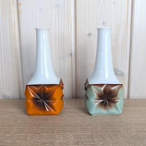 May include: Two decorative vases. Each vase has a white, elongated neck and a geometric base. One base is orange and brown, the other is light green and brown. The vases are displayed on a wooden surface.