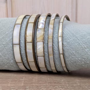 May include: Six white and gold bangles with a rectangular mother-of-pearl design. The bracelets are resting on a light blue cloth, with a wooden surface in the foreground. The bangles are a jewelry accessory.