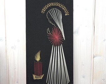 May include: A string art piece on a black background. The artwork depicts a figure with a halo, hands clasped in prayer, and a candle. The figure's dress is made of white string, and the candle and halo are gold.