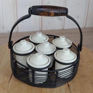 Puede incluir: Un juego de seis pequeños tarros de cerámica blanquecina con tapas, en una cesta de alambre negro con asa de madera. Los tarros tienen un borde negro y un pequeño pomo en la parte superior.