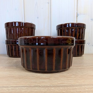Può includere: Un set di cinque pirottini in ceramica marrone. Ogni pirottino ha un esterno a coste e una finitura lucida. I pirottini sono disposti su una superficie di legno chiaro, alcuni impilati. Ideali per cuocere e servire porzioni individuali.