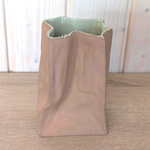 May include: A ceramic vase shaped like a crumpled paper bag with a matte light brown exterior and a smooth, pale green interior. The top edge features a distinct serrated, zigzag pattern that mimics the opening of a traditional paper sack.