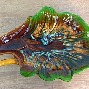 Puede incluir: Un plato de servir de cerámica con forma de hoja con un glaseado marrón, verde y amarillo. El plato tiene un asa en un lado.