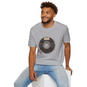 May include: A gray t-shirt featuring a vintage film reel graphic. The reel is black and gray with a label in the center. The shirt is worn by a person wearing blue jeans.