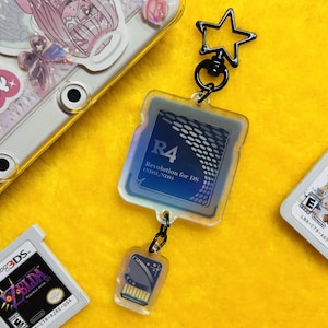 May include: A clear acrylic keychain featuring a rectangular R4 Revolution for DS game card design with a blue gradient background and white text, attached to a smaller SD card charm and a black star-shaped metal clasp ring.