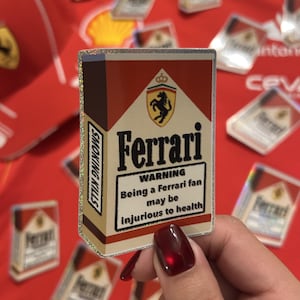 May include: A rectangular sticker with a glittery border featuring a red and white design resembling a cigarette pack. It displays the Ferrari logo, the text Ferrari, and a warning box stating: WARNING Being a Ferrari fan may be injurious to health.