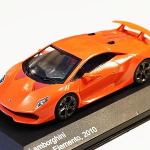 May include: Orange Lamborghini Sesto Elemento 2010 model car on a black display stand. The model car has black wheels and a rear spoiler. The display stand has the text "1 of 1.000 pcs".