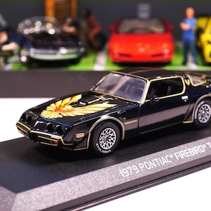 May include: A black 1979 Pontiac Firebird T/A model car with gold trim and a gold and orange eagle graphic on the hood. The model is displayed on a black base. Other toy cars and figures are in the background.