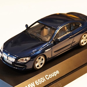 Könnte beinhalten: Ein dunkelblaues BMW 650i Coupe Modellauto. Das Miniaturfahrzeug steht auf einem schwarzen Präsentationsständer mit dem BMW-Logo und dem Text "BMW 650i Coupe". Das Auto hat silberne Räder und einen detaillierten Innenraum.