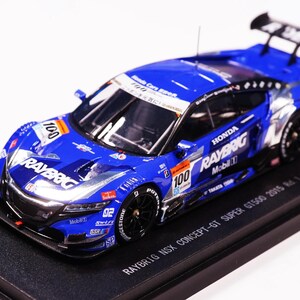Op de afbeelding: Een blauwe Raybrig Honda NSX Concept-GT Super GT500 2015 schaalmodel raceauto met nummer 100, witte sponsorstickers, zwarte wielen en een achterspoiler, gemonteerd op een zwarte rechthoekige displayvoet met gedrukte tekst op het oppervlak.