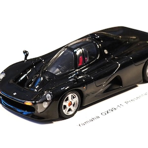 Op de afbeelding: Zwart modelauto van een Yamaha OX99-11. Het miniatuurvoertuig heeft een gestroomlijnd ontwerp met een transparante cockpit en rood interieur. De wielen hebben zilveren velgen. De tekst "Yamaha OX99-11 Presentation" is zichtbaar.