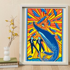 May include: A framed print of a blue whale jumping out of the water with two figures in black suits and white shirts standing in front of it. The background is a yellow and orange sunburst with smaller blue fish swimming around the whale.